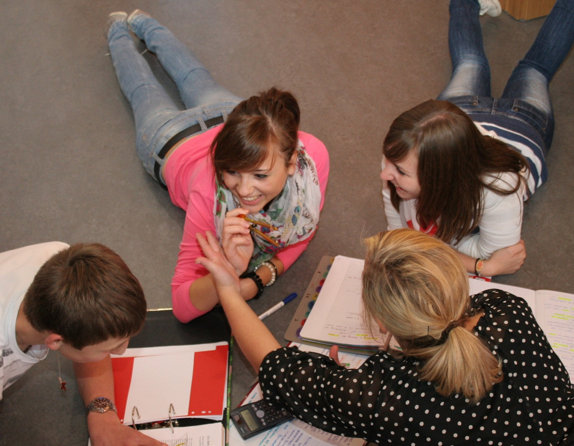 4 lachende Jugendliche liegen bei Hausaufgaben auf dem Boden